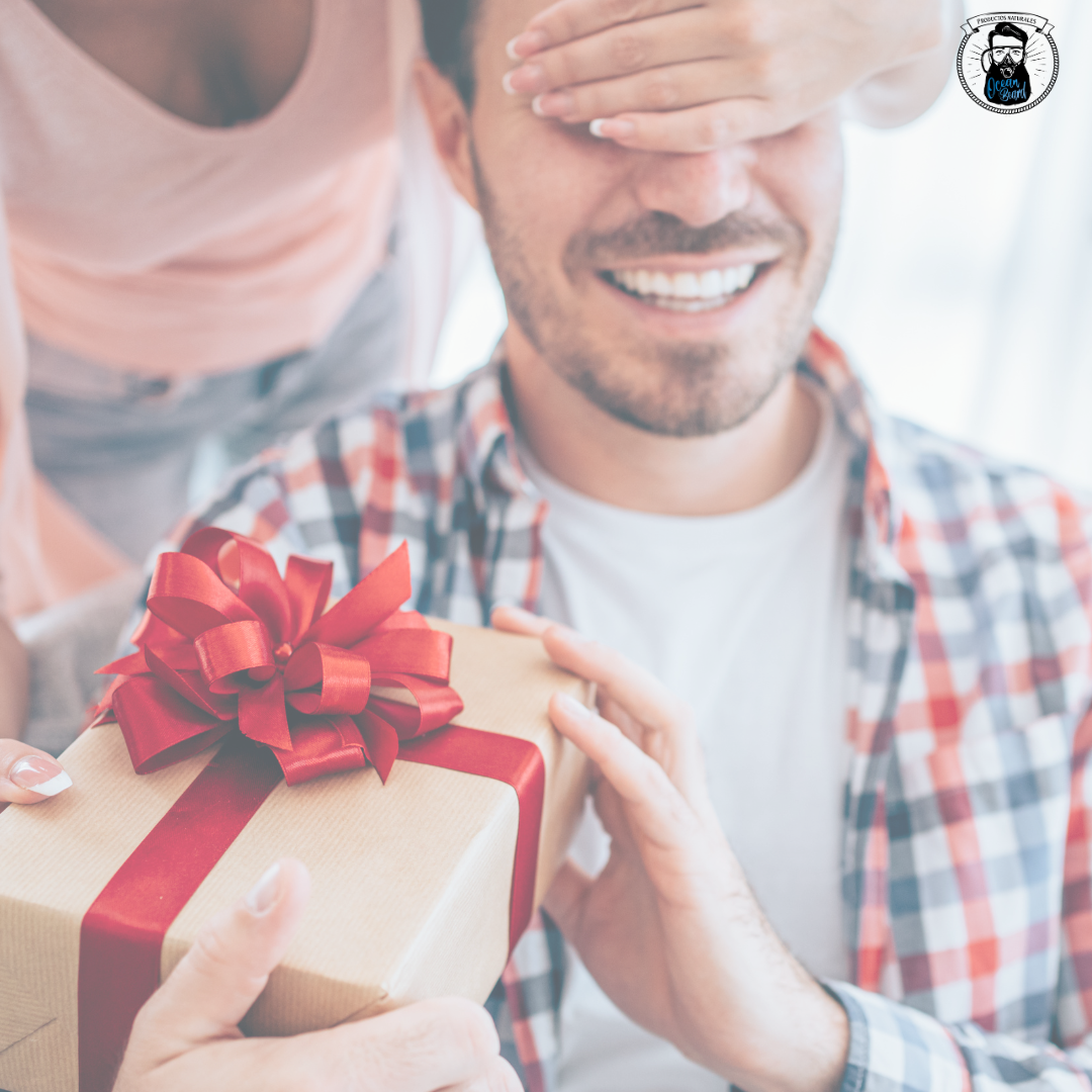 Hombre joven con los ojos cerrados y actitud expectante recibiendo un regalo de cuidado de barba de parte de una mujer